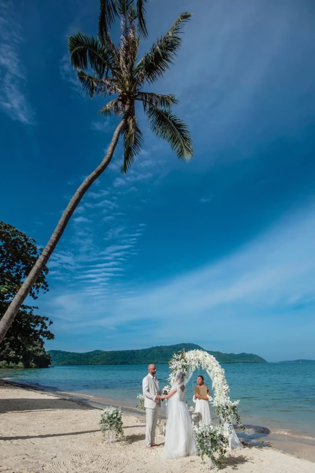 Couple exchanging vows under a floral arch on an island beach with leaning palm tree, destination wedding makeup by Elani Artistry